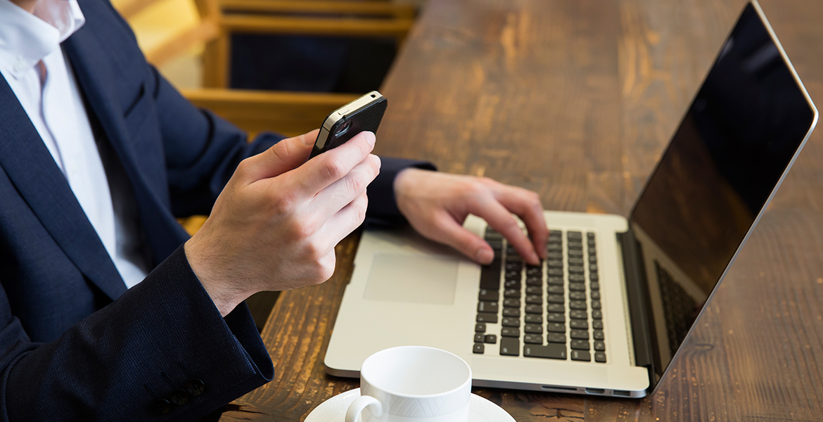 カフェでテーブルの上にノートPC、右手にスマホを持つスーツ姿の男性